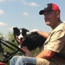 Man sitting on a tractor with a black and white dog, enjoying a sunny day outdoors. The scene captures a rural lifestyle, highlighting companionship between the man and his pet.
