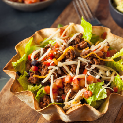 Colorful taco salad served in a crispy tortilla bowl, featuring layers of lettuce, seasoned ground beef, black beans, diced tomatoes, and shredded cheese, garnished with fresh ingredients.