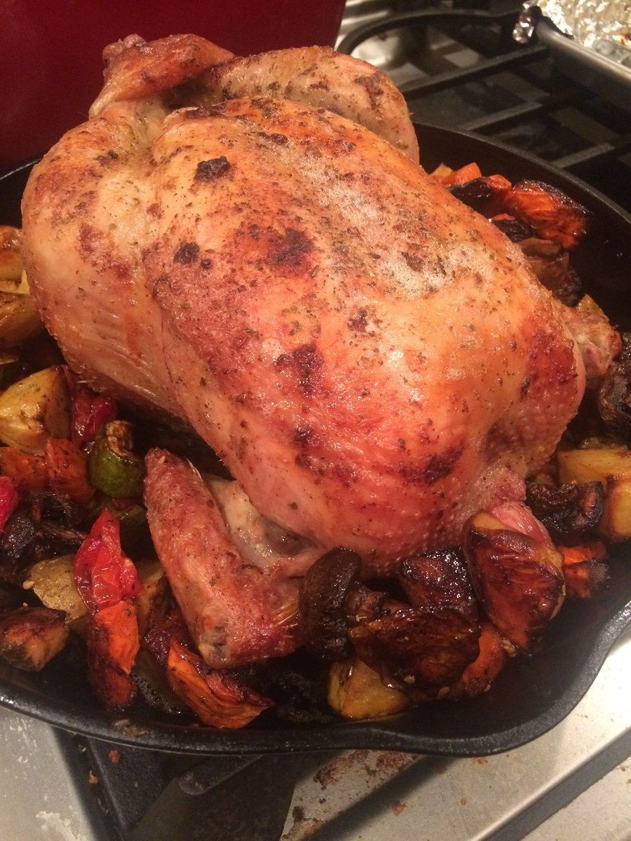 Roasted whole chicken in a cast iron skillet surrounded by colorful roasted vegetables, including potatoes, tomatoes, and carrots, showcasing a golden-brown, crispy skin.